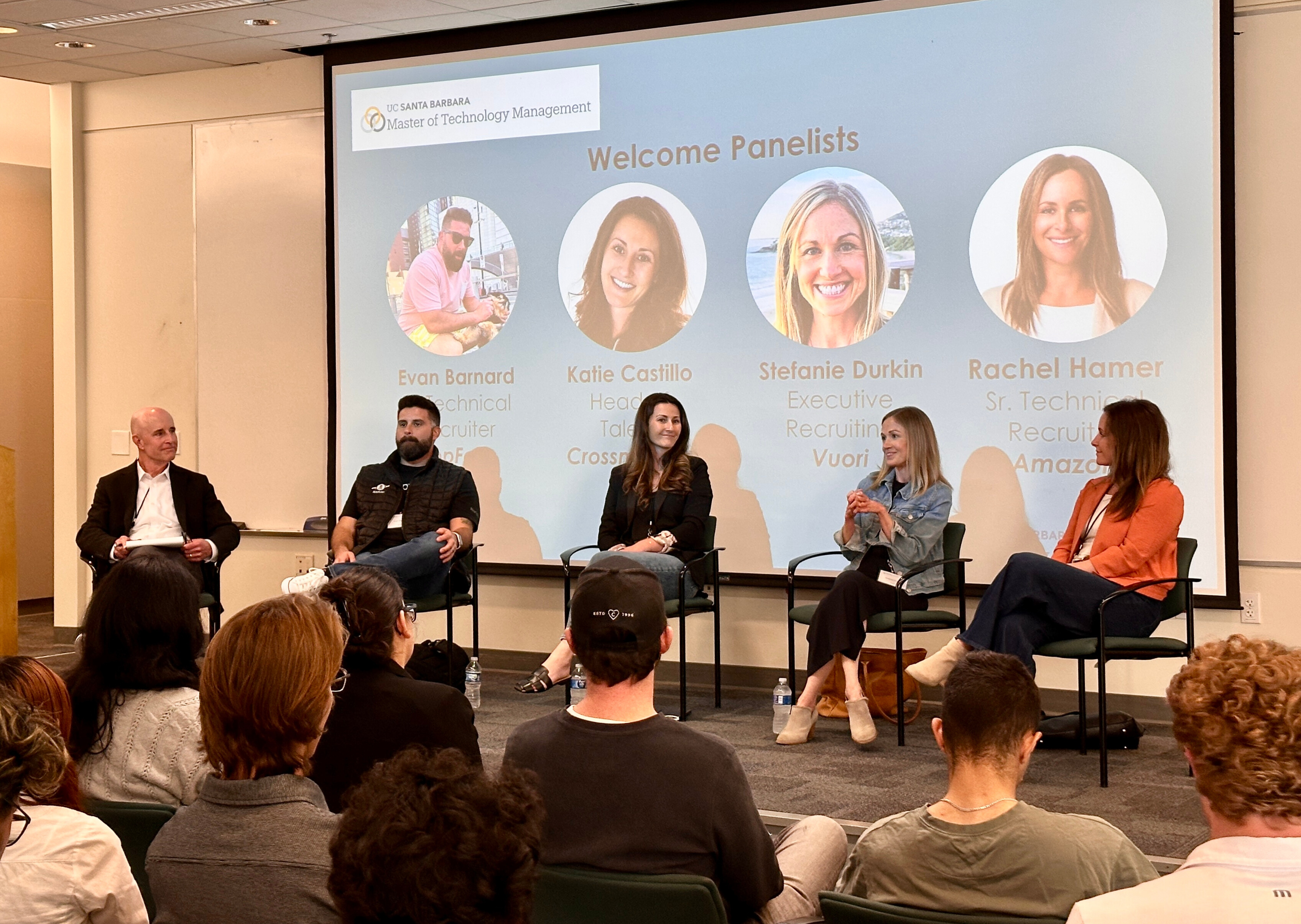 A group of 5 panelists speaking to an audience with a projector screen in the background.