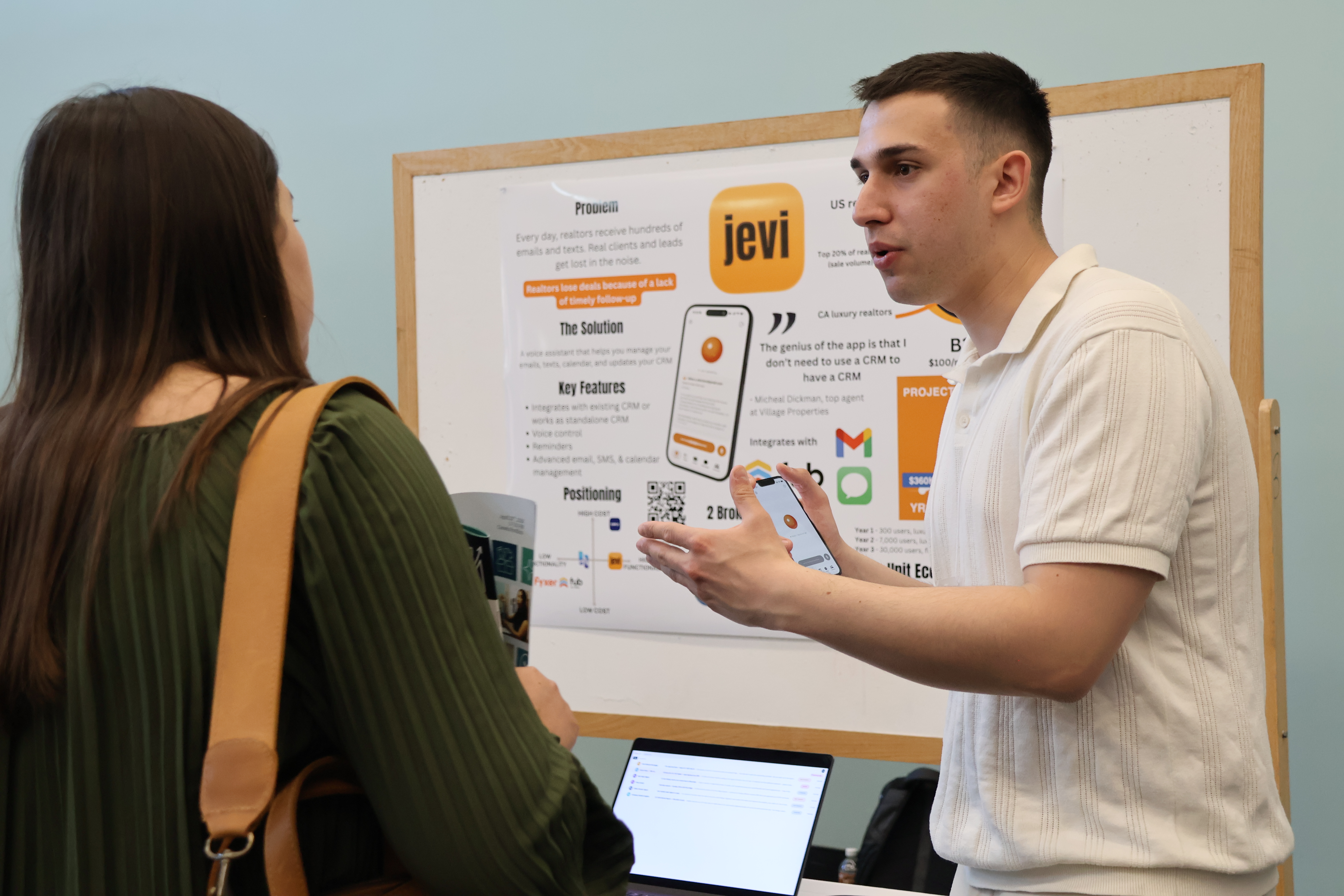 A student in front of a posterboard shows off an app on a smartphone