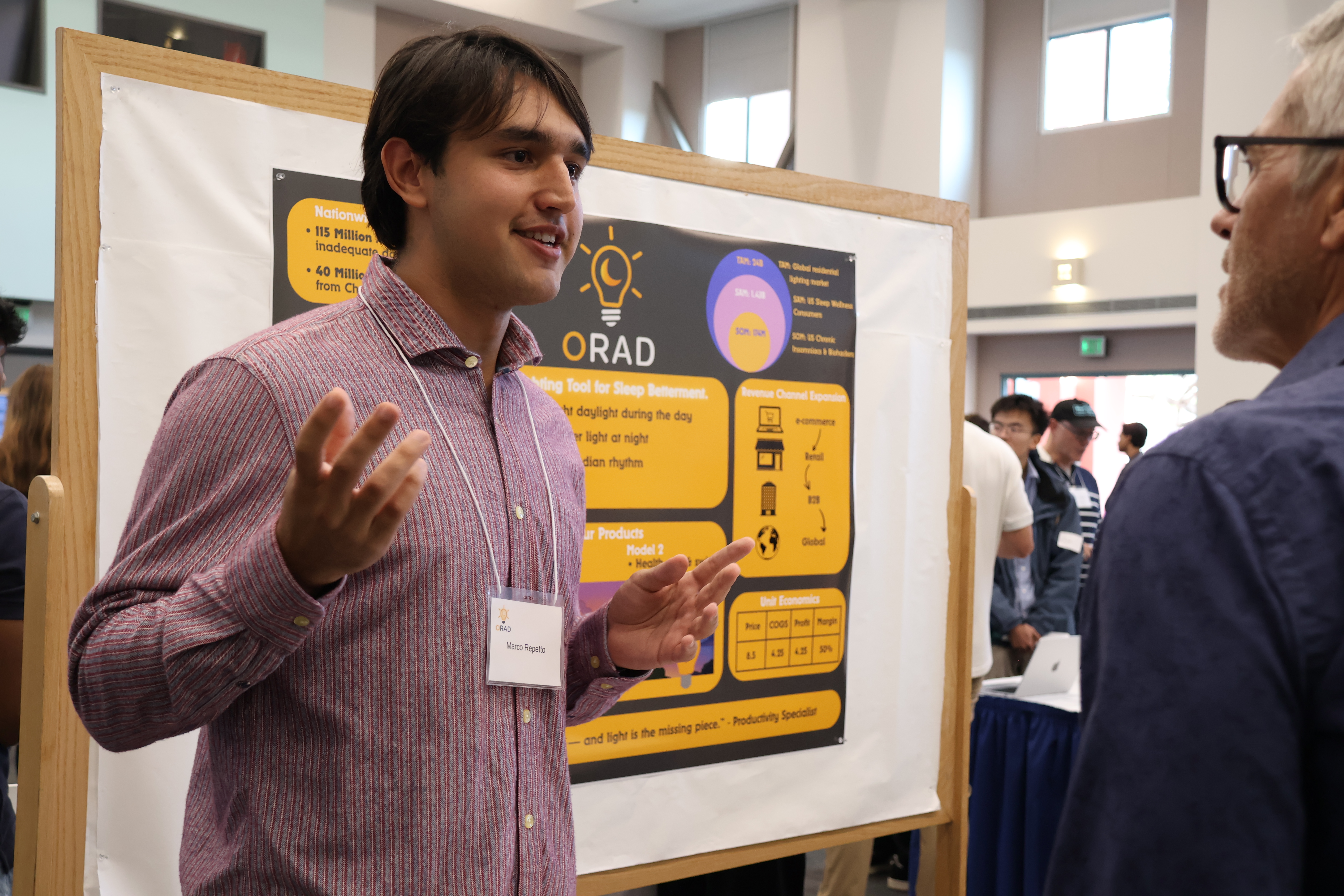 A student in front of a posterboard pitches his product to a fair attendee