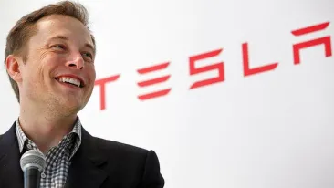 lower angle shot of elon musk with a tesla sign behind him