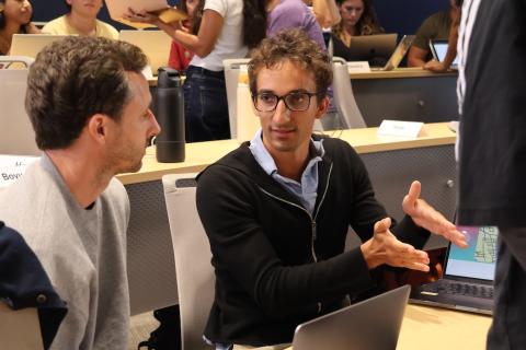 A student in a black zip up speaks to a classmate in front of a laptop.