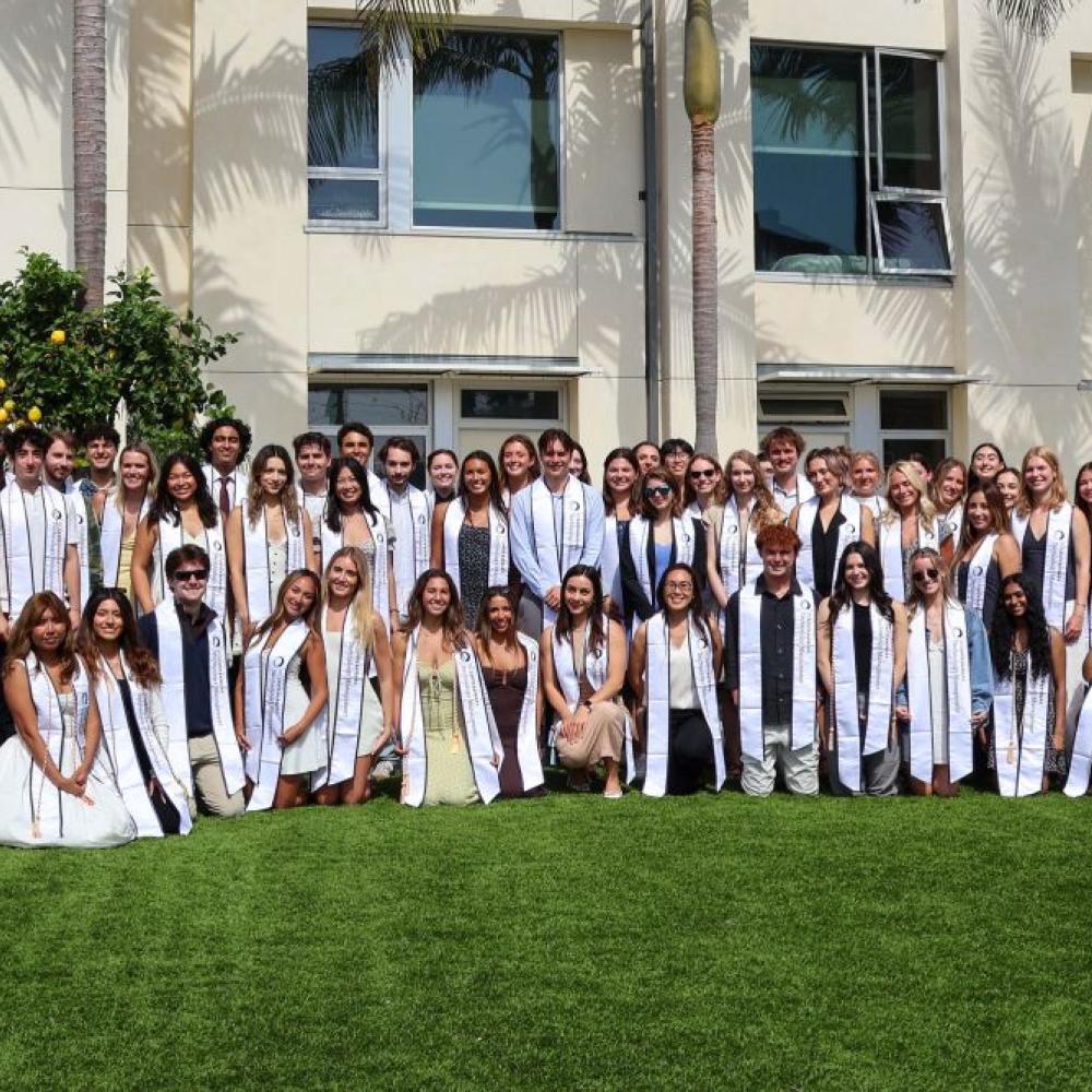 A large group of students in graduation stolls sitting and standing on a lawn outdoors