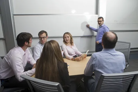 a group of businesspeople sitting at a table haveing a meeting. one of them is standing up writing on a whiteboard