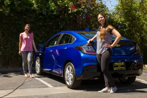 two girls posing in front of a blue car that is using a charging station
