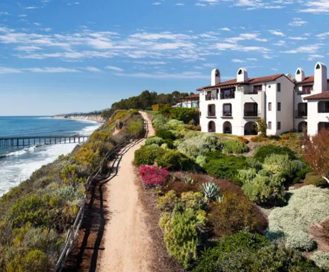 mid-arial photo of a dirt trail with the coast to its left and a white building to its right