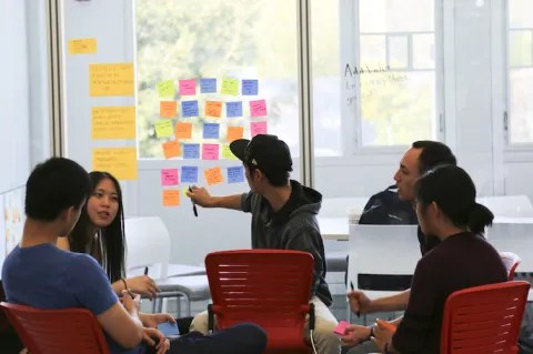 five students sit at a table in discussion. one student puts up post-it-notes on the window behind them