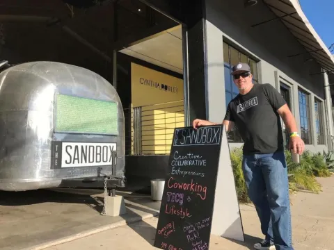 photo of a man standing next to the sandbox airstream. he is wearing a sandbox i-shirt and is leaning on a chalk menu with sandbox info on it