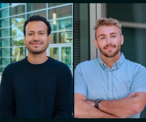 Two men, Ph.D. students in Technology Management, one wearing dark sweater, the other in a short sleeved blue shirt, are seen standing in front of a modern building at the UCSB campus. 